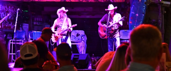 Two country singers with guitars performing on a stage lit in purple during the Pensacola Beach Songwriters Festival